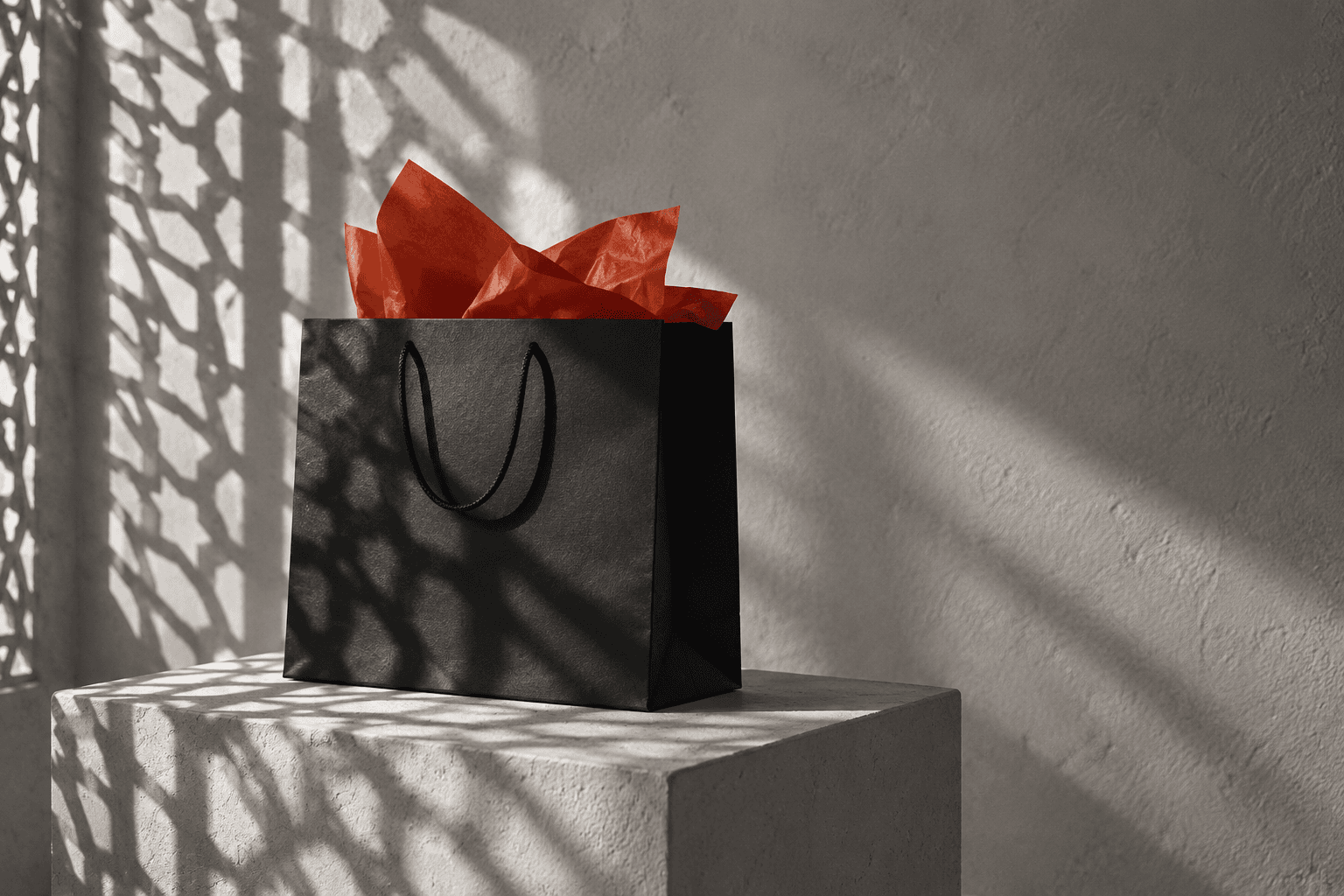 A black shopping bag with red tissue paper on a plaster plinth, under a mashrabiya shadow